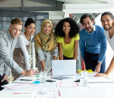 A smiling group of multiethnic students at a desk in front of a laptop looking at the camera