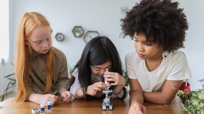 Three grade school students conducting a science experiment as one peers through a microscope.
