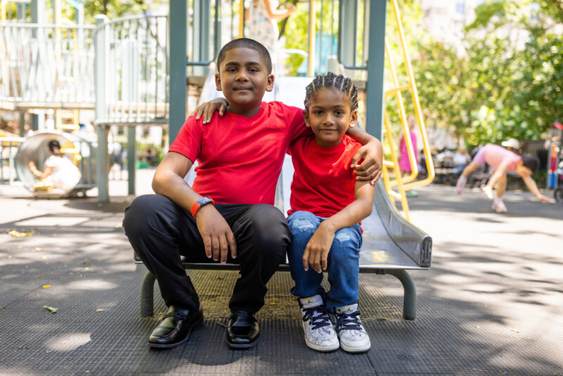 Brothers King and Messiah Singh smiling and sitting arm-in-arm on a sliding board at a park. The two were born with a rare blood desire known as G6PD and must receive regular blood transfusions.