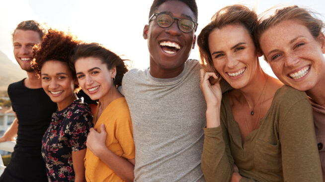 Diverse group of young adults outdoors and smiling with arms interlocked.