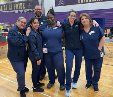 A group of six multi-ethnic New York Blood Center staff locked arm-in-arm smiling at a blood drive in a gymnasium.