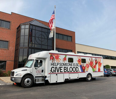 A white Rhode Island Blood Center blood drive bus with the message HELP SOMEONE ELSE. GIVE BLOOD. The bus has pastel-colored hands painted around the message.