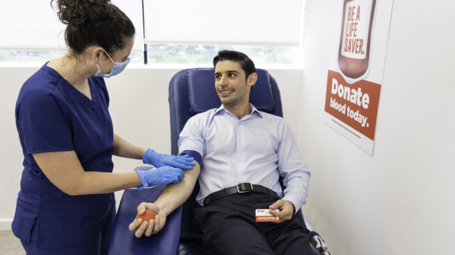 A male blood donor being prepared to donate blood by a Rhode Island Blood Center phlebotomist. A sign reading, Be a Lifesaver, Donate Blood, is in the background.