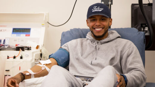 Smiling young man in a baseball cap and sweatshirt donating blood at a Connecticut Blood Center location.