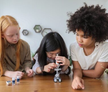 Three children conducting a science experiment with one observing a sample through a microscope between them.