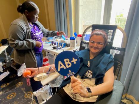 Blood donor in donation chair with blue sign reading AB+ being cared for by BBD phlebotomist.