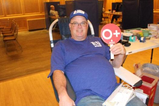 Male blood donor in donation chair smiling and holding up O+ sign.