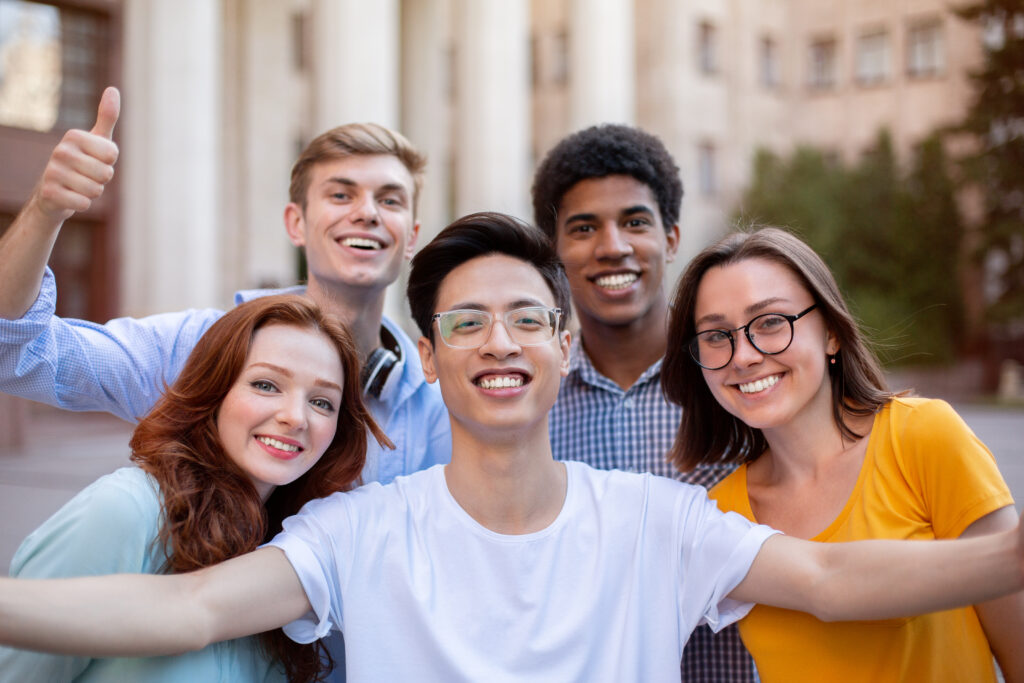 Five multi-ethnic students smiling in front of a building.