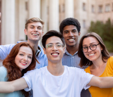 Five multi-ethnic college students smiling in front a building.